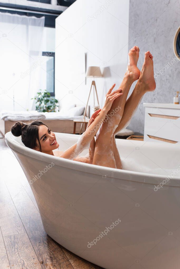 young joyful woman with raised legs taking bath in bathtub with bath foam 
