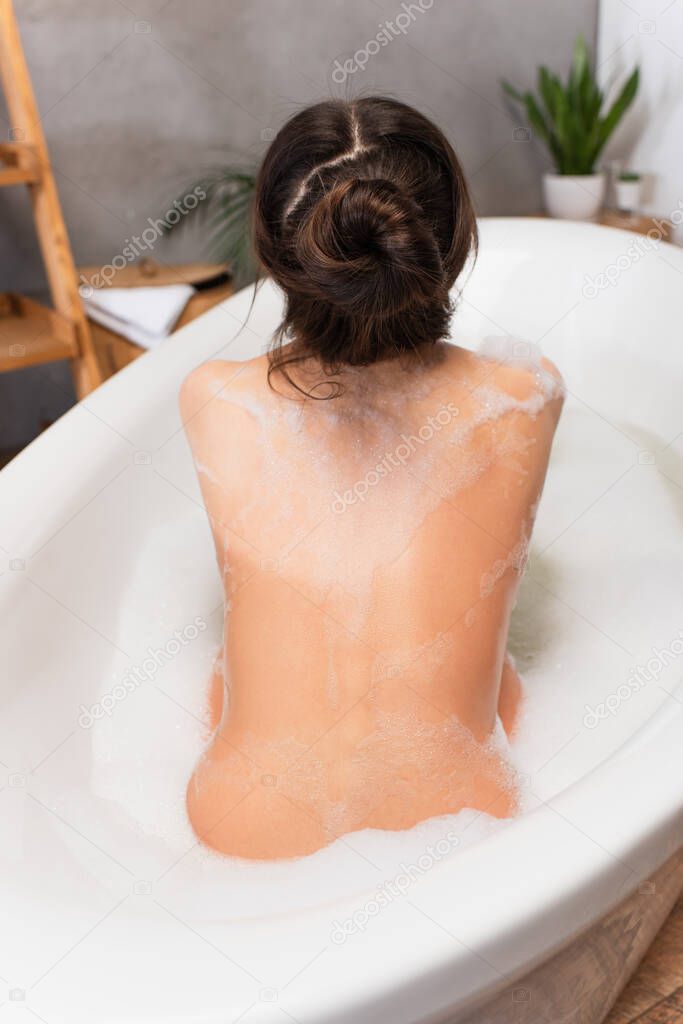back view of wet young woman sitting in bathtub with bath foam