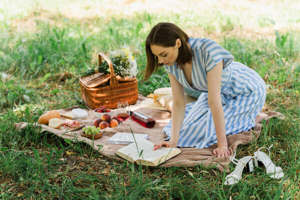 Woman in dress reading book near wine, fruits and blurred basket on blanket in park 