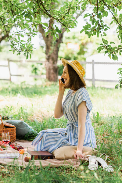 Side view of smiling woman talking on mobile phone near food and wine on blanket in park 