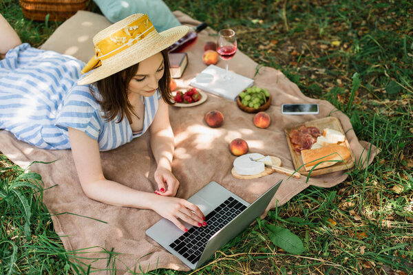 Высокоугольный вид женщины в солнцезащитной шляпе с ноутбуком рядом с едой на одеяле в парке 