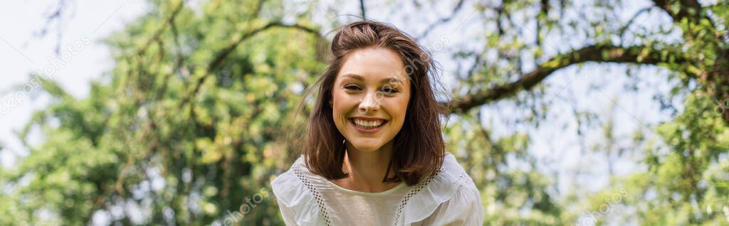 Smiling woman looking at camera in park during summer, banner 