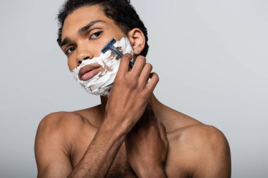 Gri renkli kameraya bakarken traş olurken güvenlik jiletiyle tıraş olan üstsüz Afrikalı Amerikalı adam.