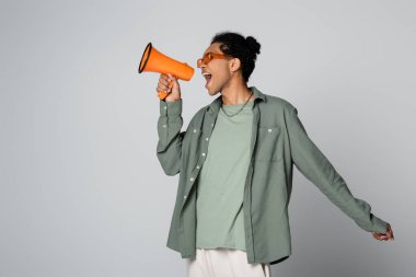 trendy african american guy shouting in orange megaphone isolated on grey clipart