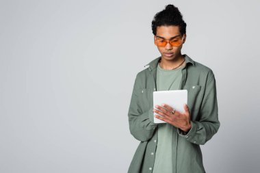Gri üzerine izole edilmiş dijital tablet kullanan şık gömlekli ve gözlüklü Afro-Amerikan genç adam.