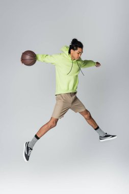Kapüşonlu, şortlu ve spor ayakkabılı, gri arka planda basketbolla atlayan Afro-Amerikan bir adam.