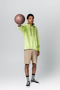 Yeşil kapüşonlu ve bej şortlu Afro-Amerikan adam gri arka planda basketbol oynuyor.