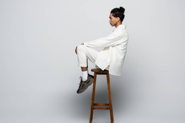 side view of african american man sitting on high stool in white suit and sneakers on grey background