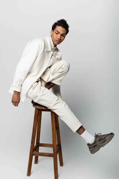 young african american man in trendy suit and sneakers posing on high stool on grey background