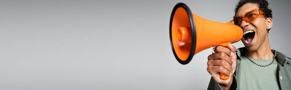 молодой африканский американец в модных очках кричит в громкоговорителе изолированном на сером, баннере