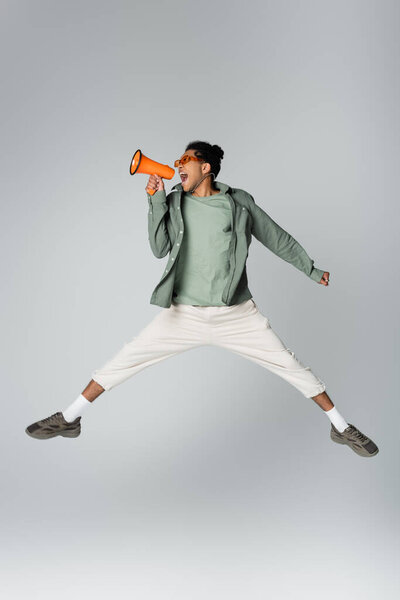 african american man in trendy clothes jumping with megaphone on grey background