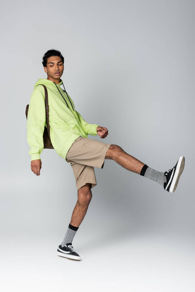 trendy african american man in hoodie and shorts posing on one leg isolated on grey