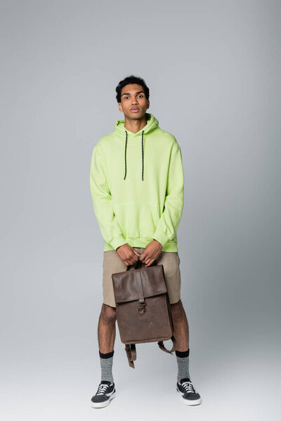 full length view of african american guy in green hoodie standing with leather backpack isolated on grey