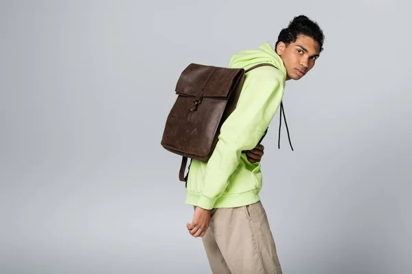 trendy african american man with brown, leather backpack looking at camera isolated on grey