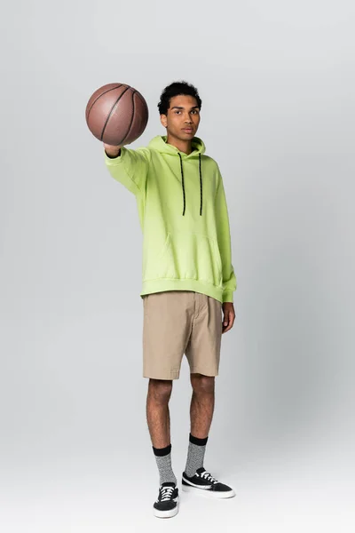 african american guy in green hoodie and beige shorts showing basketball on grey background