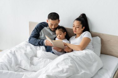 Asyalı ebeveynler yeni yürümeye başlayan kızlarına kitap okuyorlar.
