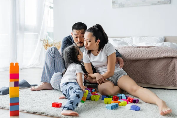 asian parents pouting lips near toddler daughter and building blocks