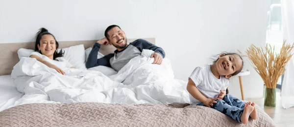 Alegre Asiático Criança Sentado Cama Perto Borrado Pais Banner — Fotografia de Stock