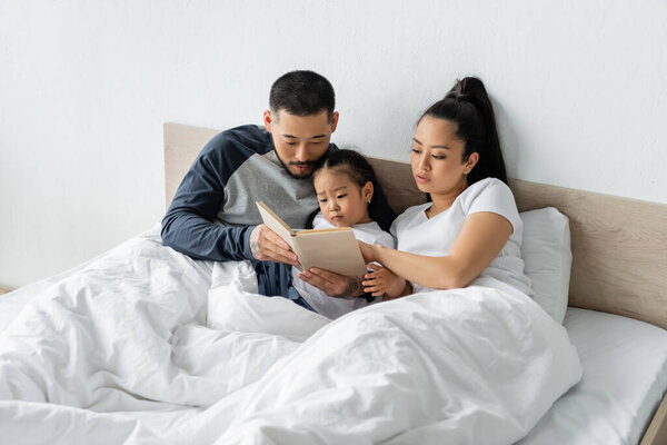 asian parents reading book to toddler daughter in bed