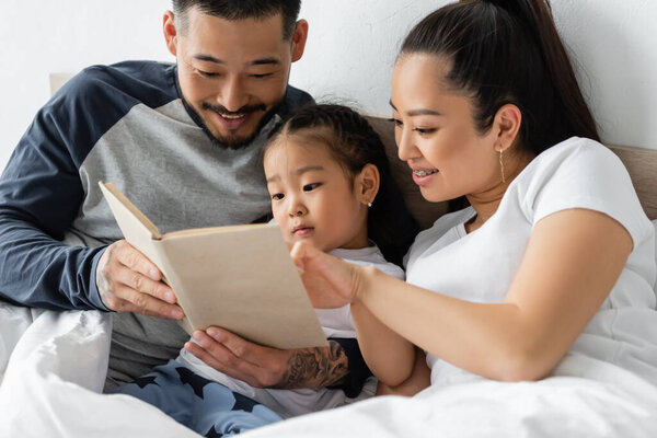 smiling asian parents reading book to toddler daughter in bed