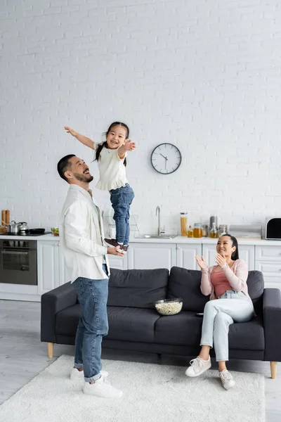 asian father holding in arms daughter near happy wife sitting on couch