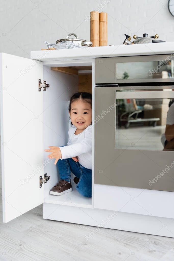 happy toddler asian kid hiding in kitchen cabinet