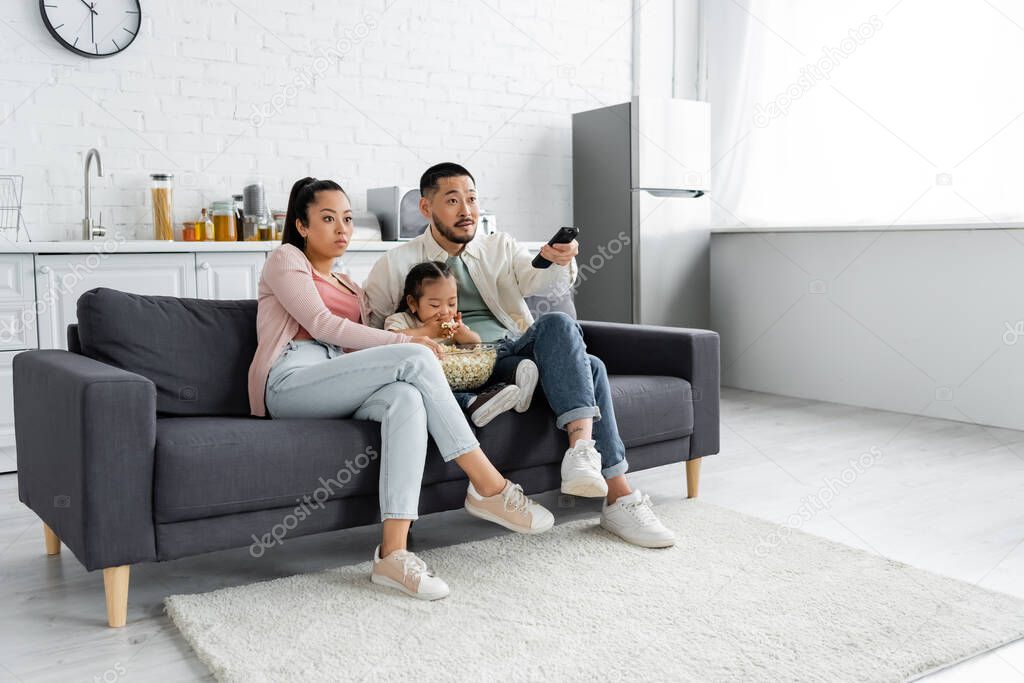 focused asian family watching movie and kid eating popcorn in living room