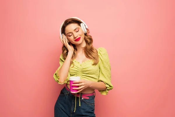 Pleased Woman Adjusting Wireless Headphones Holding Paper Cup Isolated Pink — Stock Photo, Image