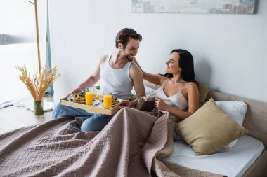 Sakallı adam kahvaltı tepsisini tutuyor ve yatakta neşeli kız arkadaşına bakıyor.