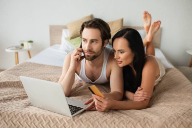 Adam elinde kredi kartı tutuyor ve kız arkadaşıyla internetten alışveriş yaparken bilgisayarın yanında akıllı telefonla konuşuyor. 