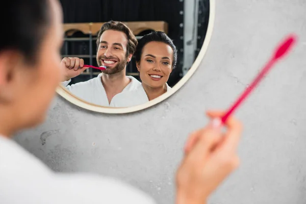 reflection in mirror of happy young couple in white bathrobes brushing teeth