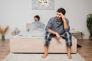 man sitting on bed and looking aside after disagreement with boyfriend on blurred background, relationship difficulties concept clipart