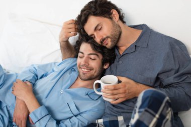 happy gay man holding cup of tea and hugging boyfriend in bed clipart