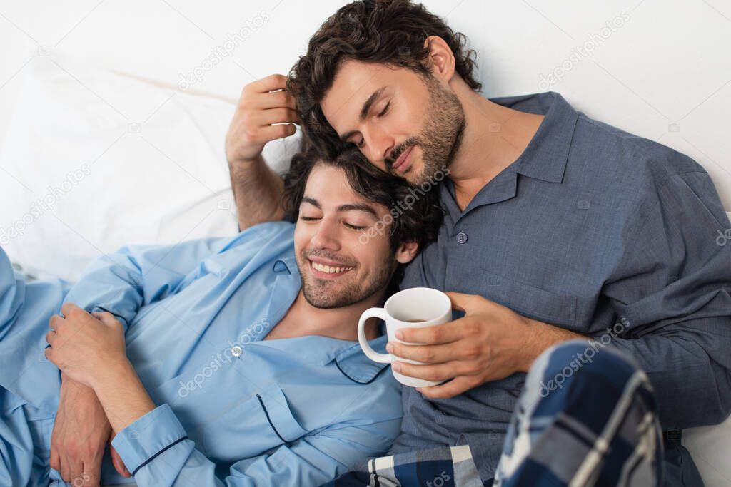 happy gay man holding cup of tea and hugging boyfriend in bed