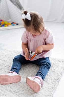 toddler girl with down syndrome sticking out tongue and playing with toy sheep and plane on carpet  clipart