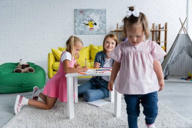 Gülümseyen anaokulu öğretmeni oyun odasında yürüyen bulanık sendromlu çocuğa bakıyor. 