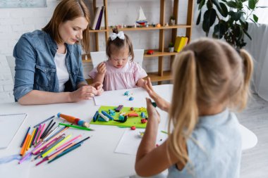 kindergarten teacher molding plasticine with disabled kid with down syndrome near blurred girl  clipart