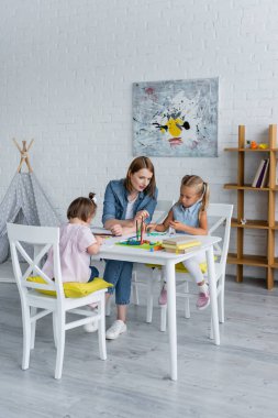 Anaokulundaki mutlu öğretmen, Down sendromlu engelli bir çocuğun yanında plastik kalıplar döşeyen anaokulu çocuğuna bakıyor.