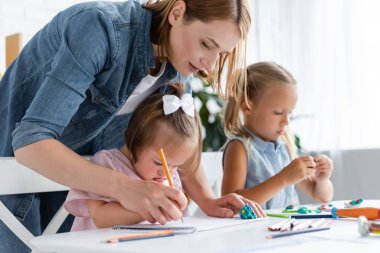 teacher assisting disabled child with down syndrome drawing near blurred child in private kindergarten  clipart
