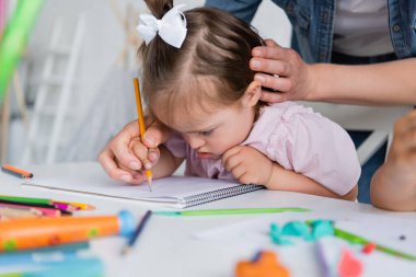 teacher assisting disabled girl with down syndrome drawing near blurred child in private kindergarten  clipart