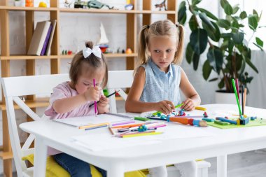 blonde girl looking at disabled toddler child with down syndrome drawing in private kindergarten  clipart
