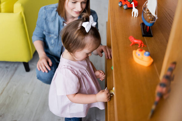 toddler girl with down syndrome looking at toys on wooden shelf near smiling kindergarten teacher