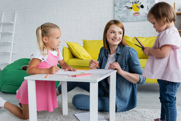 smiling kindergarten teacher and preschooler girl looking at kid with down syndrome holding color pencils 
