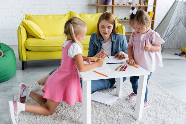 smiling kindergarten teacher talking with preschooler child and kid with down syndrome near papers and color pencils 