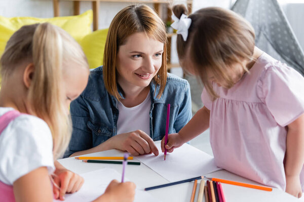 smiling kindergarten teacher looking at blurred toddler kid with down syndrome drawing with preschooler girl 