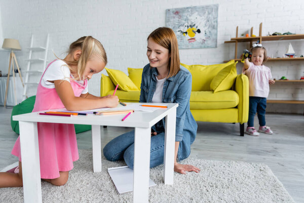счастливая воспитательница детского сада смотрит на рисунок девочки в то время как ребенок с синдромом Дауна стоит на размытом фоне