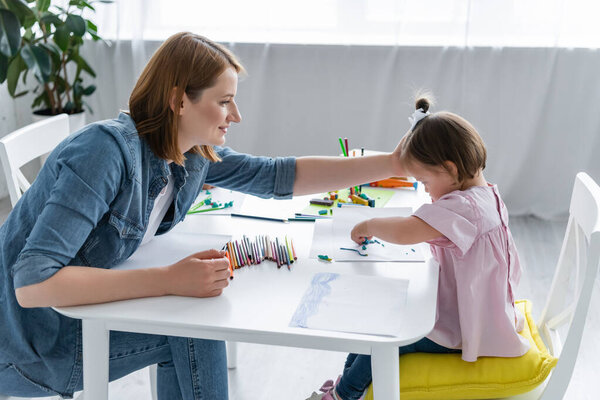 happy kindergarten teacher reaching disabled child with down syndrome molding plasticine on paper