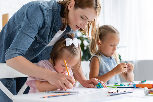 teacher assisting disabled child with down syndrome drawing near blurred child in private kindergarten 