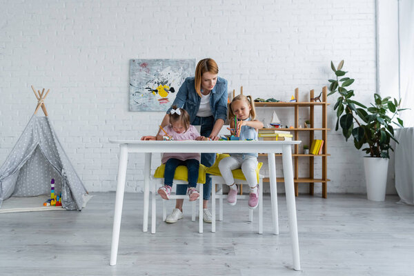 педагог, стоящий позади дошкольницы и ребенка-инвалида с синдромом Дауна рисунок в частном детском саду 