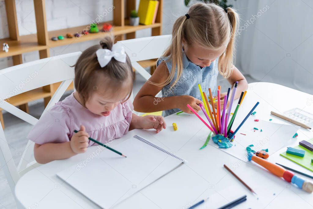 high angle view of girl with down syndrome drawing on paper near child in playroom 